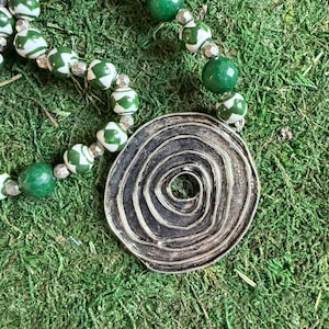 May include: A necklace featuring a large, circular, silver-toned metal pendant with a spiral design. The strand consists of alternating green jade-like beads, white beads with green patterns, and small, faceted metallic silver spacer beads.