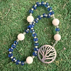May include: A beaded necklace featuring alternating faceted deep blue crystal beads and smooth pale pink round beads, accented with small silver-toned spacers. A circular silver-toned pendant with a cutout tree design hangs from the strand.