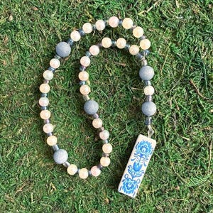 May include: A beaded necklace featuring a mix of round cream-colored pearls, translucent blue glass beads, and matte grey spherical beads. A rectangular white pendant hangs from the strand, decorated with a blue floral folk art pattern.