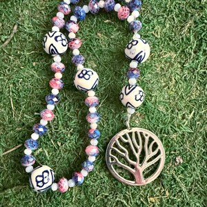 May include: A beaded necklace featuring a silver-toned circular tree pendant. The strand consists of alternating small white beads, faceted blue and pink marbled beads, and larger white ceramic beads decorated with blue floral patterns.