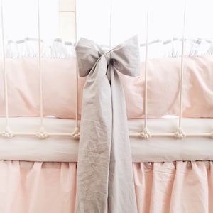 May include: A light gray bow tied around a white crib with a pink ruffled skirt. The crib is made of metal and has a white mattress.