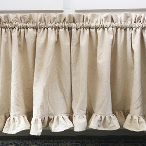 May include: A pair of off-white, ruffled kitchen valances hang from a black curtain rod. The valances are made of a lightweight fabric with a subtle texture.