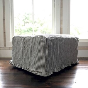 Pu&ograve; includere: Un pouf rettangolare rivestito in tessuto a righe verticali blu navy e bianche. Il tessuto ha un bordo a balze sul fondo. Il pouf &egrave; posto su un pavimento in legno scuro davanti a una finestra.