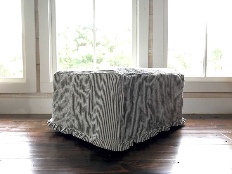 Pu&ograve; includere: Un pouf rettangolare rivestito in tessuto a righe verticali blu navy e bianche. Il tessuto ha un bordo a balze sul fondo. Il pouf &egrave; posto su un pavimento in legno scuro davanti a una finestra.