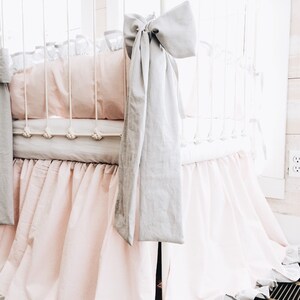 May include: A white crib with a pink and gray ruffled crib skirt. The crib is decorated with a gray bow and a white and gray patterned sheet.