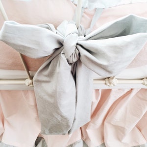 May include: A white metal bed frame with a light pink ruffled bed skirt and a gray bow tied at the top of the headboard.