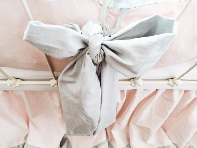 May include: A white metal bed frame with a light pink ruffled bed skirt and a gray bow tied at the top of the headboard.
