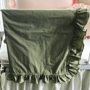 Puede incluir: Una manta de beb&eacute; de color verde salvia con un borde de volantes. La manta est&aacute; colgada de una cuna blanca.