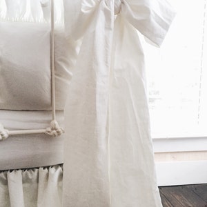 May include: White crib skirt with a large white bow tied at the top. The skirt has a ruffled edge and is made of a soft, white fabric.