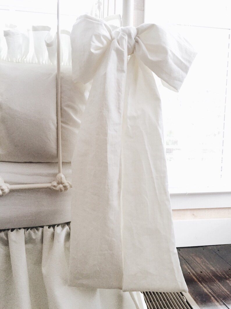 May include: White crib skirt with a large white bow tied at the top. The skirt has a ruffled edge and is made of a soft, white fabric.