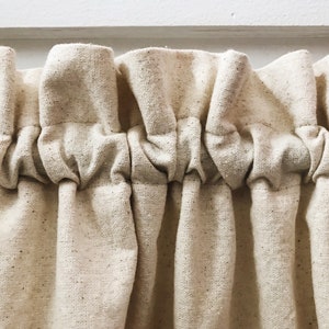 May include: Close-up of a cream-colored fabric curtain with a gathered, ruffled top. The fabric has a subtle speckled texture.