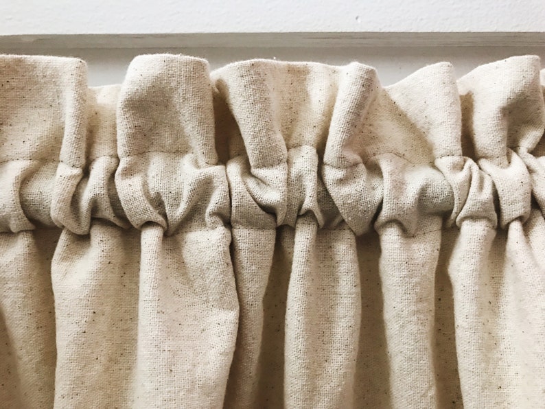 May include: Close-up of a cream-colored fabric curtain with a gathered, ruffled top. The fabric has a subtle speckled texture.