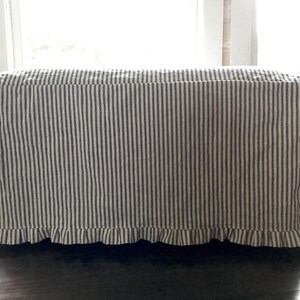 Pu&ograve; includere: Un pouf rettangolare rivestito in tessuto a righe verticali blu navy e bianche. Il bordo inferiore presenta un bordo a balze. Il pouf &egrave; posto davanti a una finestra.