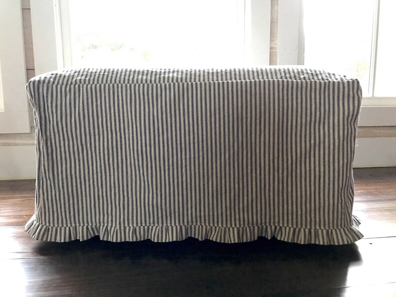 Pu&ograve; includere: Un pouf rettangolare rivestito in tessuto a righe verticali blu navy e bianche. Il bordo inferiore presenta un bordo a balze. Il pouf &egrave; posto davanti a una finestra.