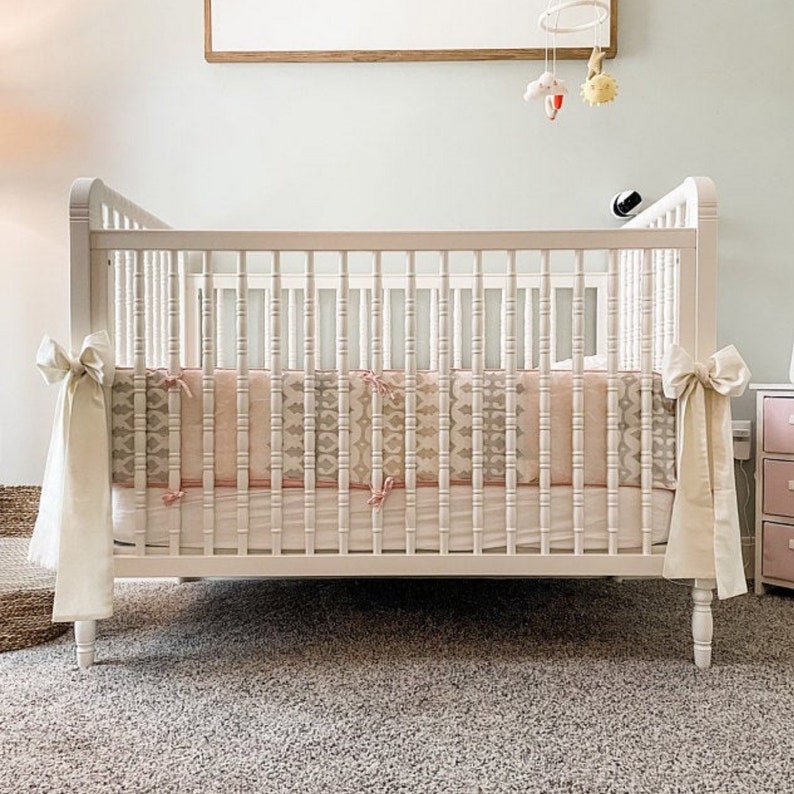 May include: White wooden crib with a white and pink patterned crib bumper and white crib skirt. The crib is on a beige carpet.