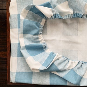 May include: A blue and white checkered fitted sheet with elastic around the edges. The sheet is folded in half and is lying on a wooden surface.