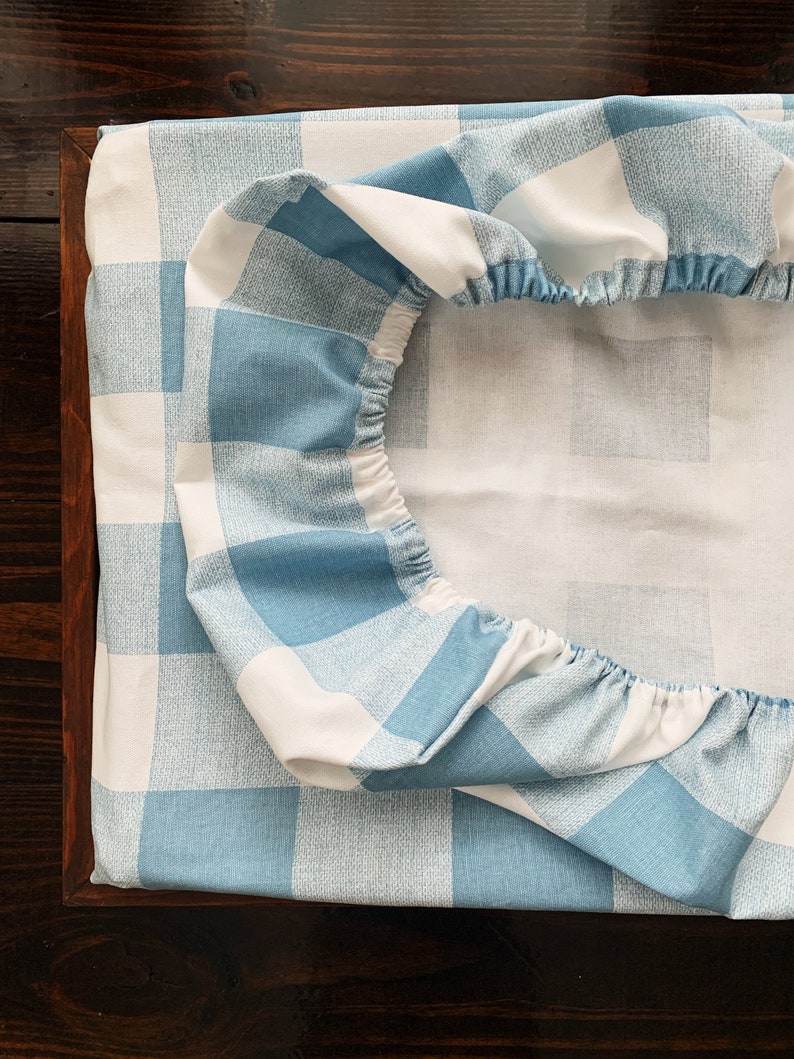 May include: A blue and white checkered fitted sheet with elastic around the edges. The sheet is folded in half and is lying on a wooden surface.