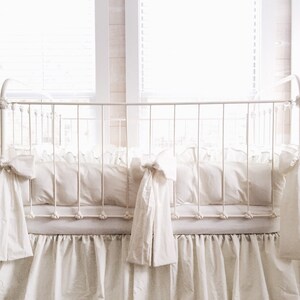 May include: White crib bedding set with a ruffled skirt and bow ties. The bedding is made of soft, white fabric and is perfect for a nursery.