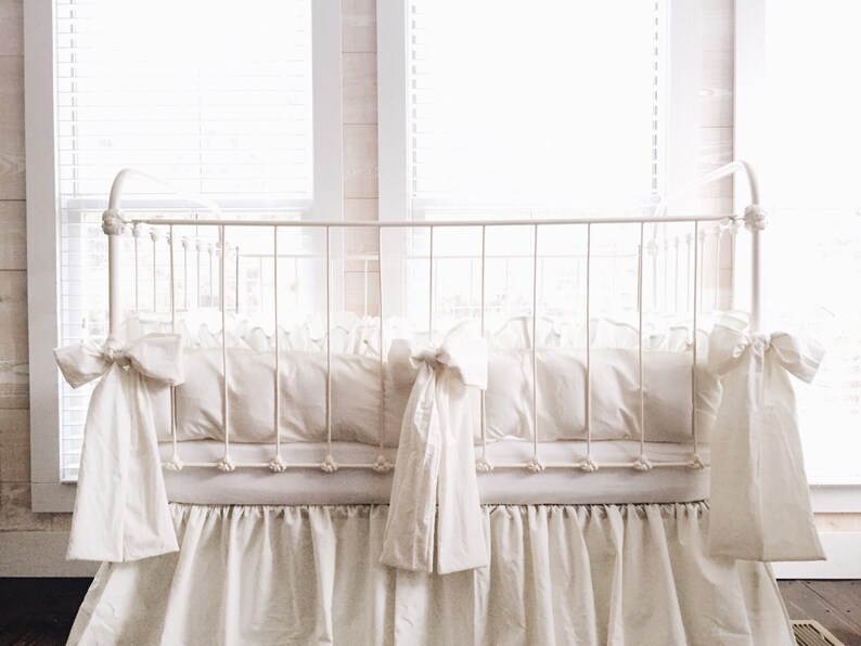 May include: White crib bedding set with a ruffled skirt and bow ties. The bedding is made of soft, white fabric and is perfect for a nursery.