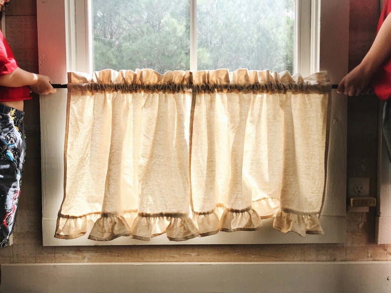 May include: Two beige ruffled curtains hang in a window with a wooden frame. The curtains are held up by a black rod. The curtains are made of a light, airy fabric.