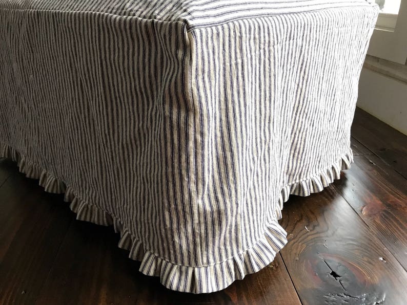 Pu&ograve; includere: Un pouf rettangolare rivestito in tessuto a righe verticali blu navy e bianco sporco. Il bordo inferiore &egrave; rifinito con un bordo a balze. Il pouf poggia su un pavimento in legno scuro.