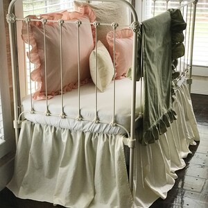 Puede incluir: Cuna de metal blanco con una falda de cama blanca con volantes, una almohada rosa con volantes blancos, una almohada blanca con ribete de encaje y una manta verde con volantes blancos.