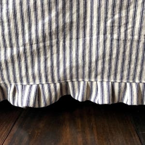 Pu&ograve; includere: Primo piano di una gonna da letto con un motivo a righe blu e bianche. Il tessuto presenta strisce verticali e un bordo a balze. La gonna &egrave; realizzata in un materiale strutturato e lo sfondo &egrave; una superficie in legno scuro.