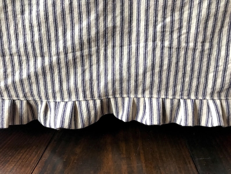 Pu&ograve; includere: Primo piano di una gonna da letto con un motivo a righe blu e bianche. Il tessuto presenta strisce verticali e un bordo a balze. La gonna &egrave; realizzata in un materiale strutturato e lo sfondo &egrave; una superficie in legno scuro.