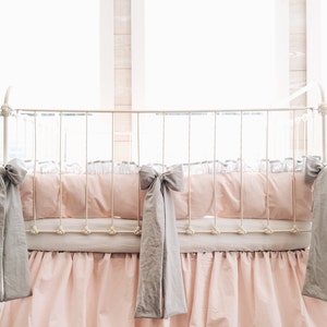 May include: A white metal crib with a pink and white ruffled crib skirt and gray bow ties. The crib is decorated with a pink and white bedding set.