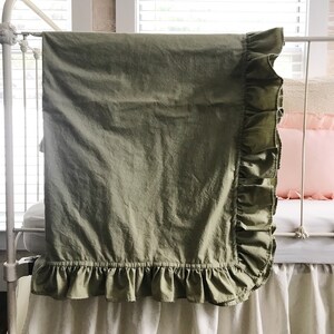 Puede incluir: Una manta de cuna verde salvia con un borde de volantes. La manta est&aacute; drapeada sobre una cuna blanca.
