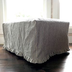 Pu&ograve; includere: Un pouf quadrato rivestito in un tessuto a righe verticali blu navy e crema. Il bordo inferiore della fodera ha un bordo a balze. Il pouf &egrave; appoggiato su un pavimento in legno scuro, con una finestra luminosa sullo sfondo.