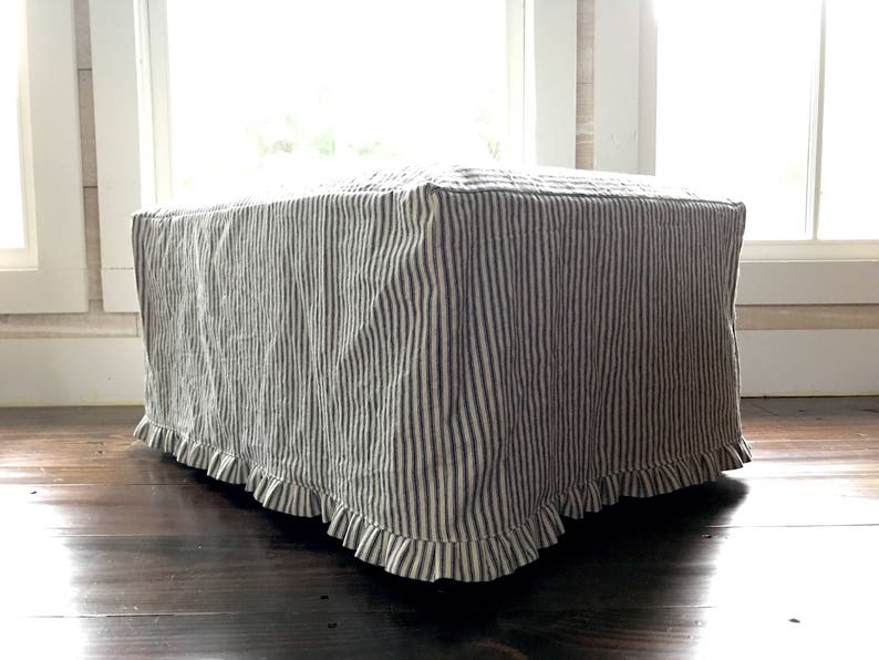 Pu&ograve; includere: Un pouf quadrato rivestito in un tessuto a righe verticali blu navy e crema. Il bordo inferiore della fodera ha un bordo a balze. Il pouf &egrave; appoggiato su un pavimento in legno scuro, con una finestra luminosa sullo sfondo.