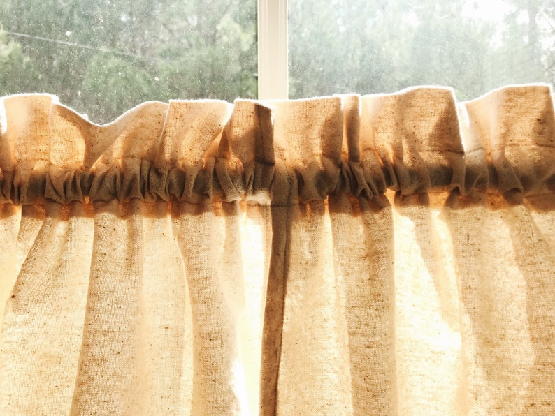 May include: A pair of off-white cotton cafe curtains with a gathered top and a simple, rustic look.