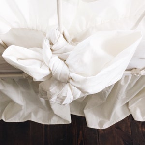 May include: White bed skirt with a ruffled edge and a white fabric bow tied at the center.