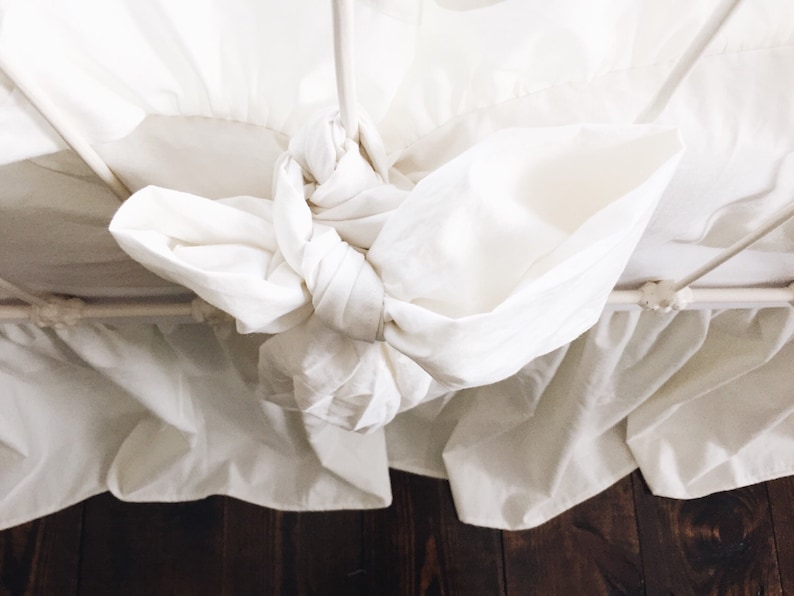 May include: White bed skirt with a ruffled edge and a white fabric bow tied at the center.