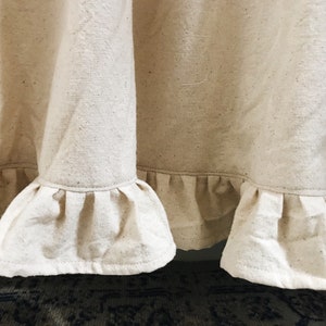 May include: A close-up of a cream-colored fabric curtain with a ruffled hem. The fabric has a subtle texture and is slightly wrinkled.