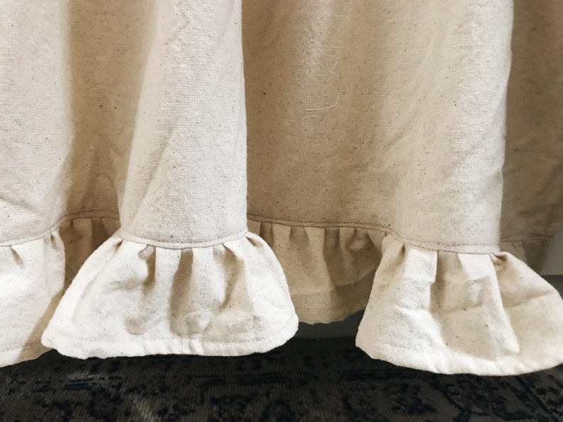 May include: A close-up of a cream-colored fabric curtain with a ruffled hem. The fabric has a subtle texture and is slightly wrinkled.