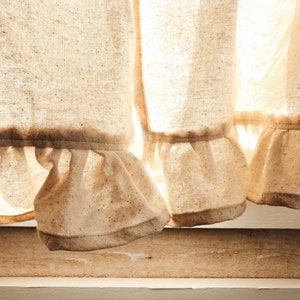 May include: Close-up of a pair of white linen curtains with a ruffled hem. The curtains are slightly sheer and allow light to shine through.