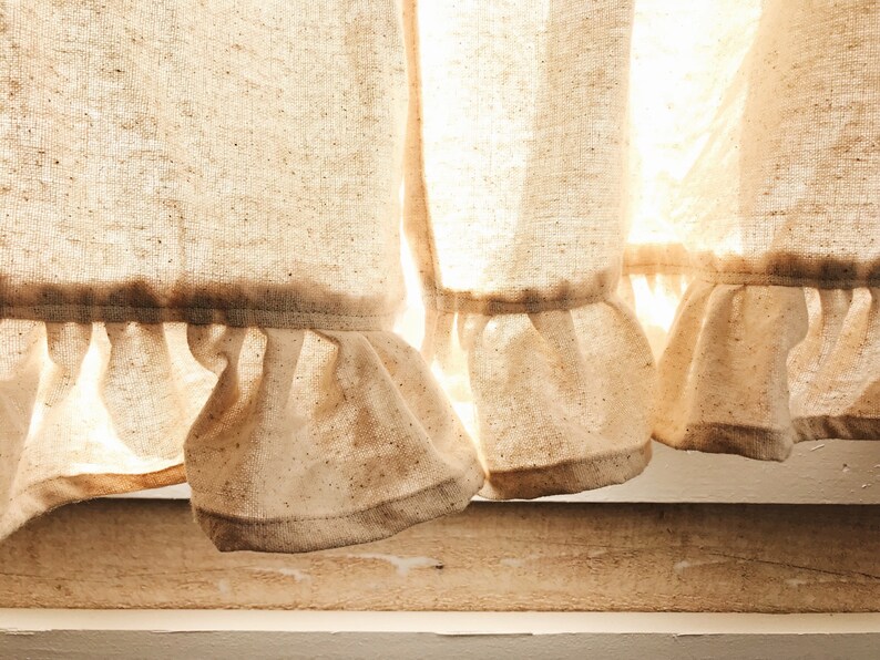 May include: Close-up of a pair of white linen curtains with a ruffled hem. The curtains are slightly sheer and allow light to shine through.