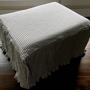 Pu&ograve; includere: Un pouf rettangolare rivestito in tessuto a righe verticali blu navy e bianche. Il tessuto ha una gonna a balze intorno alla base. Il pouf &egrave; posto su un pavimento in legno scuro.