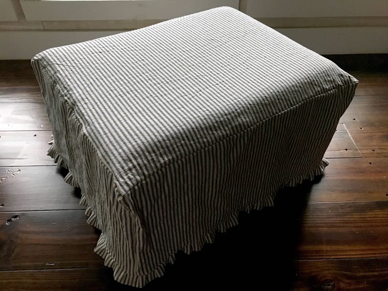 Pu&ograve; includere: Un pouf rettangolare rivestito in tessuto a righe verticali blu navy e bianche. Il tessuto ha una gonna a balze intorno alla base. Il pouf &egrave; posto su un pavimento in legno scuro.