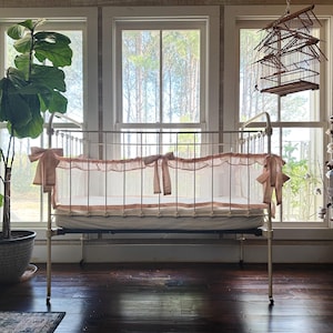 May include: A vintage-style baby crib with a metal frame and a soft, neutral-toned fabric canopy. The crib is adorned with delicate bows and set against a backdrop of large windows, a potted plant, and a decorative birdcage.