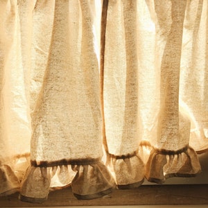 May include: A set of off-white linen curtains with a ruffled hem. The curtains are gathered at the top and hang in soft folds. Sunlight is shining through the fabric, creating a warm glow.