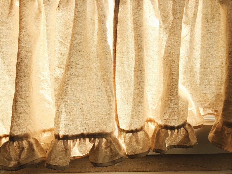 May include: A set of off-white linen curtains with a ruffled hem. The curtains are gathered at the top and hang in soft folds. Sunlight is shining through the fabric, creating a warm glow.
