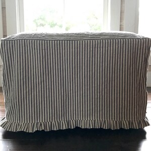Pu&ograve; includere: Un pouf rettangolare rivestito in tessuto a righe verticali blu navy e crema. Il bordo inferiore presenta un bordo a balze. Il pouf &egrave; posto su un pavimento in legno scuro, con una finestra sullo sfondo.