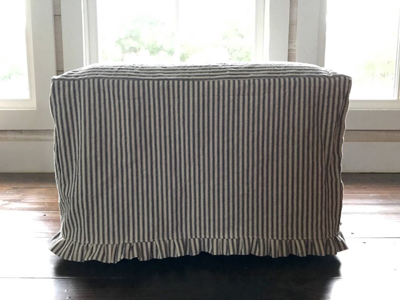 Pu&ograve; includere: Un pouf rettangolare rivestito in tessuto a righe verticali blu navy e crema. Il bordo inferiore presenta un bordo a balze. Il pouf &egrave; posto su un pavimento in legno scuro, con una finestra sullo sfondo.