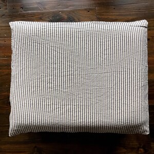 Pu&ograve; includere: Cuscino rettangolare con un motivo a righe verticali blu navy e bianco. Il cuscino &egrave; posizionato su una superficie di legno scuro, evidenziando il suo design e la sua consistenza. Il tessuto appare leggermente stropicciato.