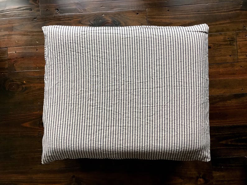 Pu&ograve; includere: Cuscino rettangolare con un motivo a righe verticali blu navy e bianco. Il cuscino &egrave; posizionato su una superficie di legno scuro, evidenziando il suo design e la sua consistenza. Il tessuto appare leggermente stropicciato.