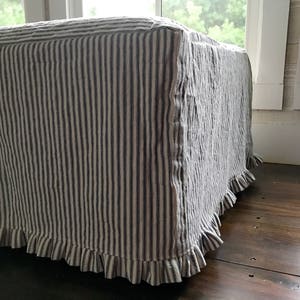 Pu&ograve; includere: Un pouf rettangolare rivestito in tessuto a righe verticali blu e bianche. Il bordo inferiore del pouf &egrave; rifinito con una gonna a balze, che aggiunge un tocco decorativo. Il pouf &egrave; posto su un pavimento in legno scuro.