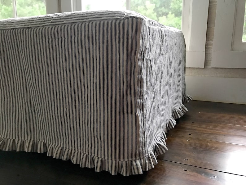 Pu&ograve; includere: Un pouf rettangolare rivestito in tessuto a righe verticali blu e bianche. Il bordo inferiore del pouf &egrave; rifinito con una gonna a balze, che aggiunge un tocco decorativo. Il pouf &egrave; posto su un pavimento in legno scuro.